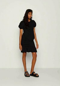Vestido preto com mangas bufantes, corpete ajustado e barra com pregas. Combinado com sandálias pretas com tachas prateadas. Tecido suave e leve.