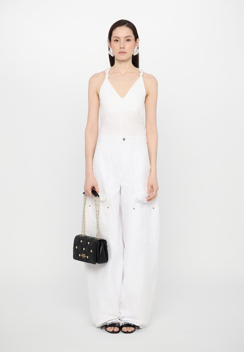 Top bianco senza maniche con scollo a V e nodi laterali, abbinato a pantaloni bianchi larghi. La modella tiene una borsa nera trapuntata con tracolla a catena.