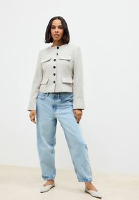Lichtgrijze cropped jack met zwarte knopen en voorzakken, gecombineerd met losse blauwe jeans en beige instappers. Zachte textuur.