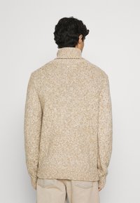 Pull beige en tricot avec un col montant, présentant une texture épaisse et une coupe légèrement ample. Aucun motif ou accent visible.