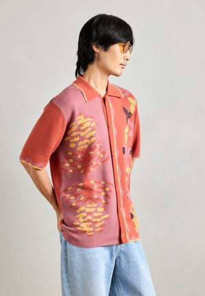 Polo à manches courtes en tricot de couleur corail et rose avec des accents jaunes. Présente des motifs abstraits de fleurs et de papillons, et une fermeture à boutons.