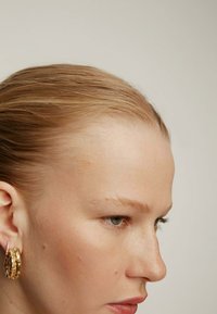 Pendientes de aro dorados con un diseño texturizado, que presentan acentos transparentes. Vista acercada del perfil de una modelo con cabello lacio y tez clara.
