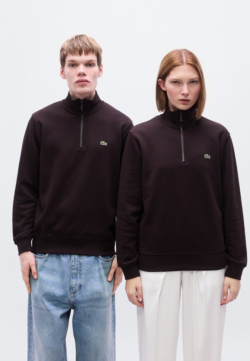 Sweat-shirts à fermeture éclair marron foncé avec col montant, ornés de petits logos verts sur le côté gauche de la poitrine, portés par-dessus un jean et des pantalons clairs.