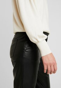 Maglione leggero di colore crema, lavorato a maglia con maniche a palloncino, abbinato a pantaloni in pelle nera aderenti che mostrano un dettaglio della tasca laterale.