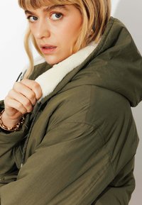 Veste vert olive avec un col en peluche blanc, fermeture éclair à l'avant et design matelassé. Le mannequin tient le col, mettant en valeur le matériau et la texture.