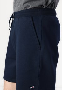 Pantaloncini blu navy con vita elastica e coulisse, tasche laterali, indossati con top grigio, piccolo logo rettangolare vicino all'orlo.