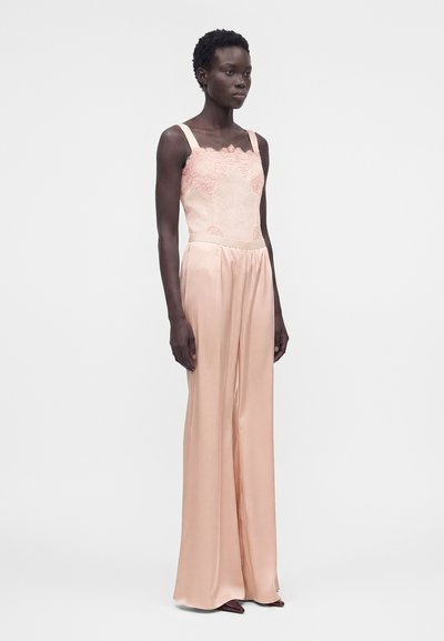 Ψηλή γυναίκα με κοντά μαλλιά που φοράει ανοιχτό ροζ κορσέ από δαντέλα και φαρδιά σατέν παντελόνια, στέκεται μπροστά σε ένα απλό φωτεινό φόντο.