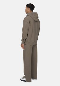 Felpa marrone con cappuccio ampio, vestibilità morbida, polsini a costine, abbinata a pantaloni larghi dello stesso colore. Design minimalista senza motivi visibili.