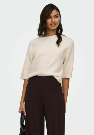 Nő krémszínű rövid ujjú pulóvert és magas derekú sötétbarna nadrágot visel, oldalán fekete kézitáskát tart.