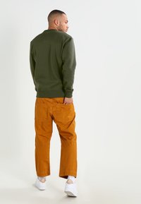 Felpa verde oliva con polsini a coste, abbinata a pantaloni cargo arancioni con tasche a toppa. Calzature: sneakers bianche con dettagli.