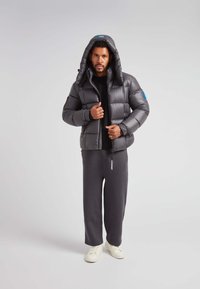 Polstret sort jakke med hætte, lynlås og blå logo-detaljer; kombineret med grå sweatpants og hvide sneakers. Glat tekstur.