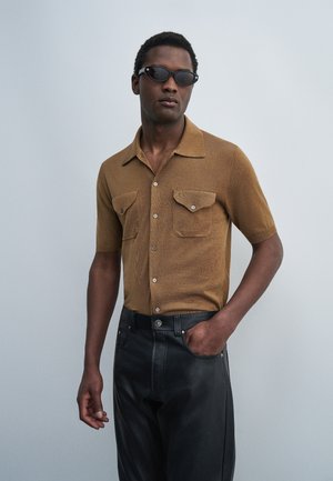Mand iført brun kortærmet skjorte med knapper og brystlommer, sorte bukser og sorte solbriller, stående med den ene hånd i lommen.