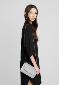 Borsa a busta con paillettes argento, chiusura a patta, tracolla e finitura testurizzata. Tenuta da una persona in un vestito nero su uno sfondo neutro.