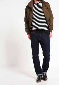 Veste aviateur kaki portée sur un pull rayé noir et blanc, associée à un jean en denim foncé et des bottes en cuir noir. Texture lisse, coupe décontractée.