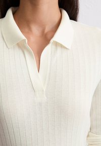Cremfarbendes, geripptes Poloshirt mit weicher Textur, das einen klassischen Kragen und eine teilweise Knopfleiste aufweist.