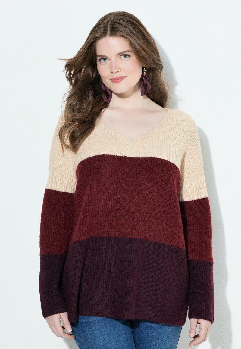 Frau mit langen braunen Haaren, die lila Ohrringe trägt und einen mehrfarbigen Pullover in Beige, Burgunderrot und Dunkelpurpur mit blauen Jeans kombiniert.