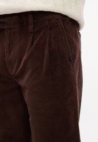 Brune kordfløjspants med en tekstureret overflade, der har folder og et lille sort logo-tag på lommen. Stoffet er blødt og ribbet.