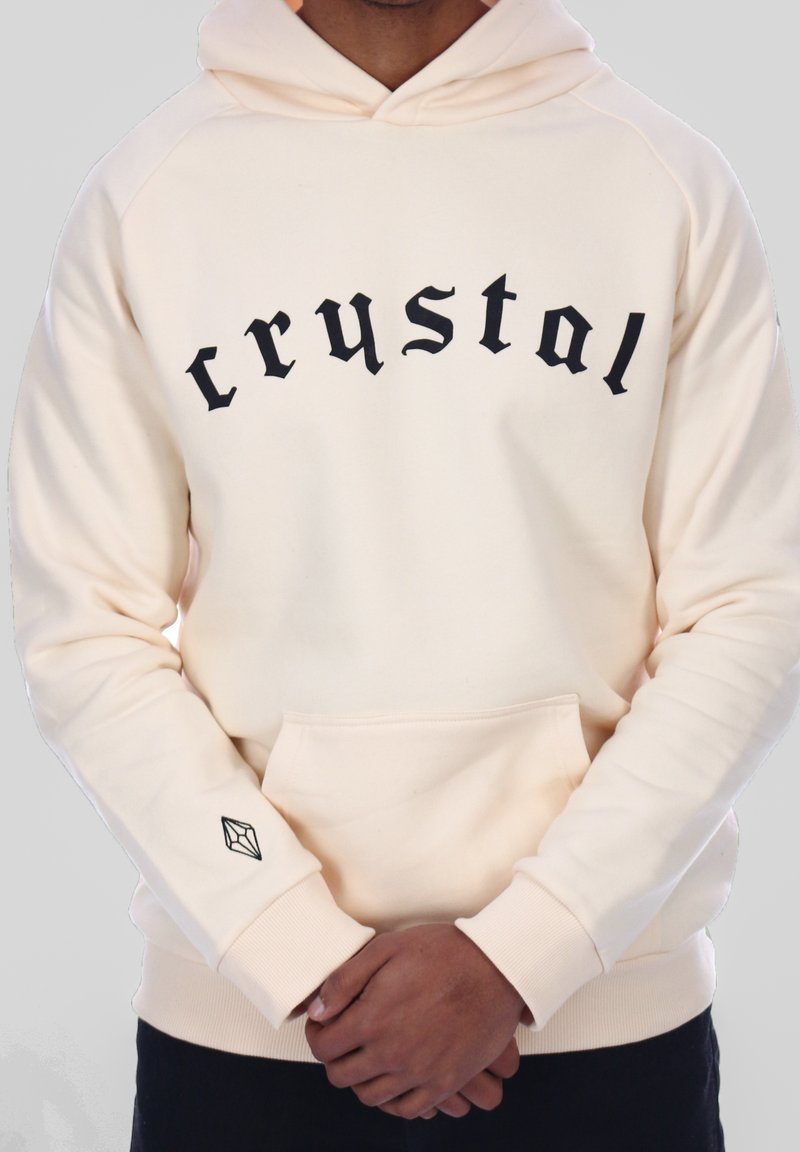 Person iført en cremefarvet hættetrøje med "crystal" i sort gotisk skrifttype på brystet og et lille geometrisk logo på venstre håndledsområde.