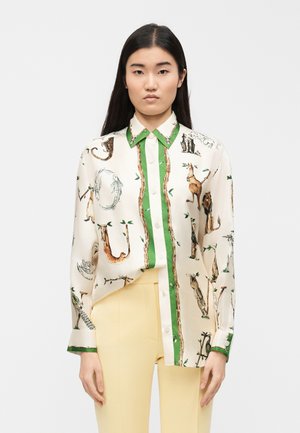 Žena v krémové blůze se zeleným lemováním a ilustracemi zvířat, v kombinaci s bledě žlutými kalhotami, stojící před jednobarevným bílým pozadím.