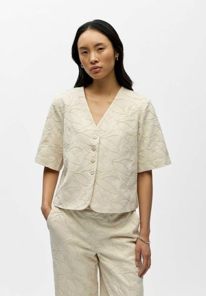 Kobieta o długich ciemnych włosach, ubrana w kremowy, krótki rękaw, zapinany na guziki top oraz pasujące spodnie z fakturą liści, stoi z rękami w kieszeniach.