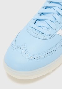 Lyseblå lærsnøre med brogue-lignende perforeringer, matchende snørebånd og en hvit aksentstripe på siden. Teksturert overflatefinish.