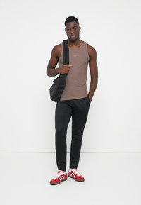 Top fără mâneci de culoare maronie, pantaloni negri și adidași roșii și albi. Modelul poartă o geantă neagră pe umărul unul. Design simplu, ajustat.