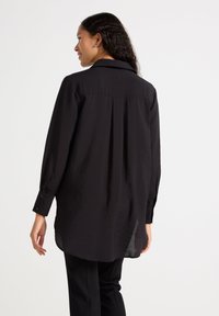 Camicia nera a maniche lunghe realizzata in un tessuto stropicciato, con colletto, polsini con bottone e orlo leggermente curvato sul retro.