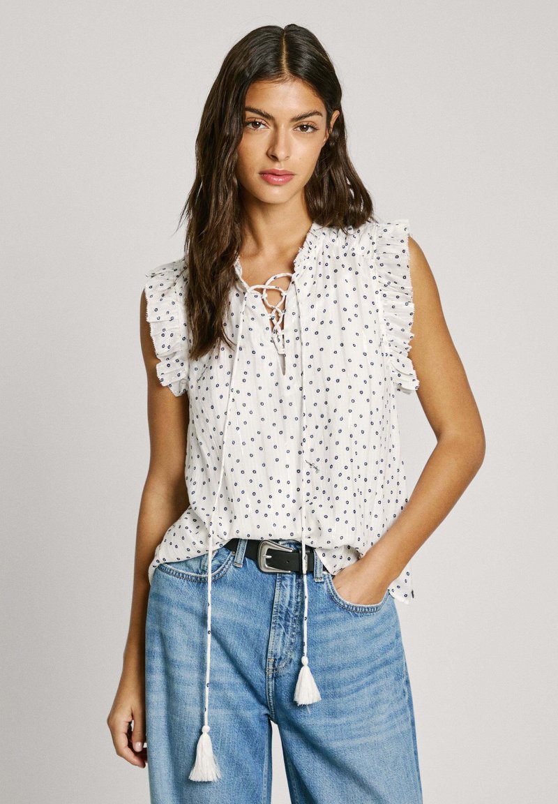 Blusa bianca senza maniche con pois blu navy, maniche arricciate e scollo con lacci e ponpon, abbinata a jeans blu a vita alta.
