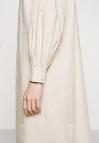 Gros plan d'une main à côté d'un vêtement beige à manches longues avec des poignets boutonnés et un tissu froncé au niveau du poignet.