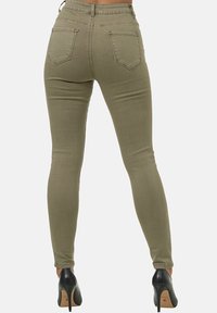 Jeans ajustados verde oliva con una textura suave, cinco bolsillos, lazos para el cinturón y un ajuste ceñido, combinados con zapatos de tacón negro.