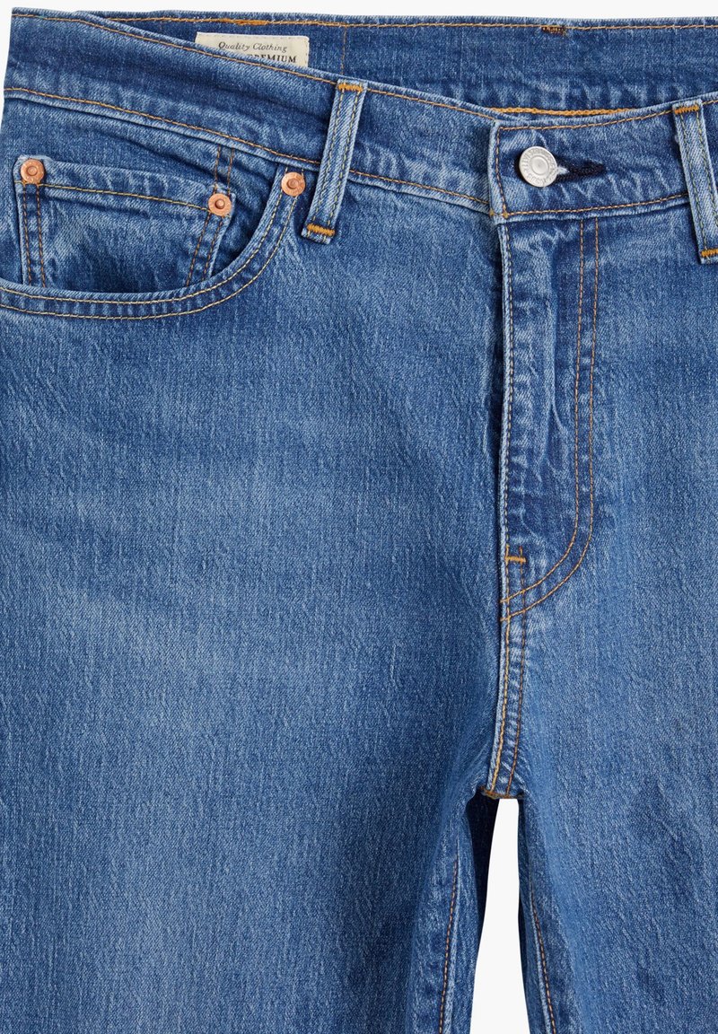 Jeans en denim bleu moyen avec des coutures orange, design à cinq poches et fermeture à bouton. Présente une étiquette de marque sur la taille.