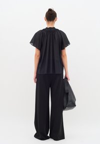 Blusa nera trasparente con colletto arricciato, maniche corte, abbinata a pantaloni neri a gamba larga. La modella tiene in mano una borsa tote nera con texture.