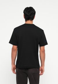 T-shirt en coton noir à manches courtes, col rond, avec un détail de logo bleu sur la manche gauche. Texture lisse et coupe régulière.