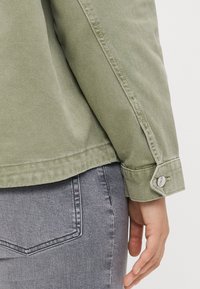 Olive green denim jacket with visible stitching, buttoned cuffs, and a textured finish, paired with gray jeans, showing a rear view.