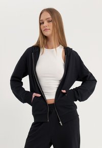 Haut de survêtement zippé noir en tissu doux, avec une coupe décontractée, des poches avant et des poignets côtelés. Porté sur un débardeur blanc.