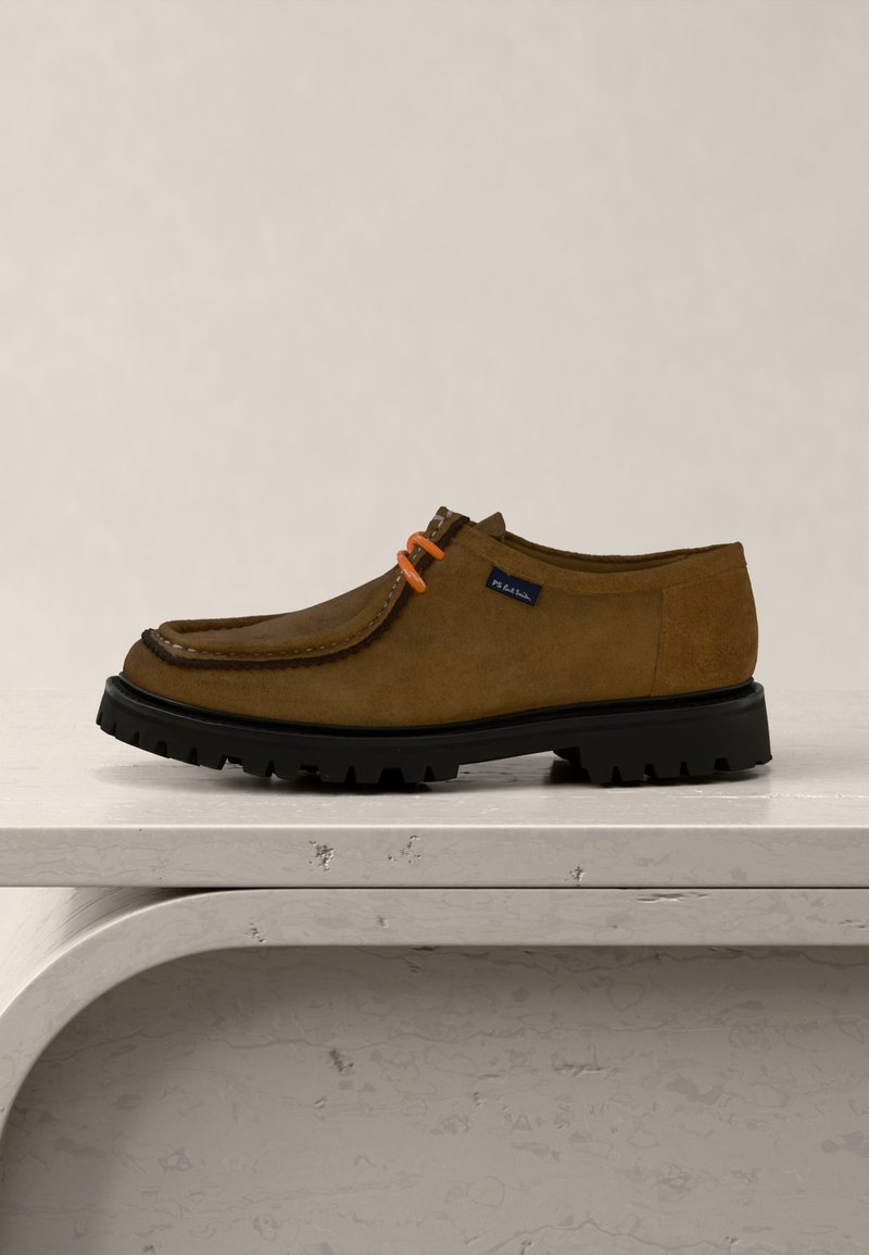 Chaussure pour homme en daim marron avec une semelle noire épaisse, des lacets orange et une petite étiquette « PS Paul Smith » sur le côté, présentée sur une surface blanche texturée.