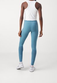 Niebieskie legginsy sportowe wykonane z elastycznego materiału. Posiadają wysoki pas i kontrastowe panele. Noszone z białym bezrękawnikiem i białymi tenisówkami.