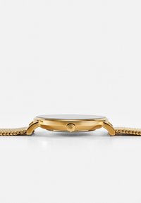 Montre analogique dorée avec un cadran noir rond, un boîtier élancé et un bracelet en maille métallique texturée avec une fermeture à cliquet.