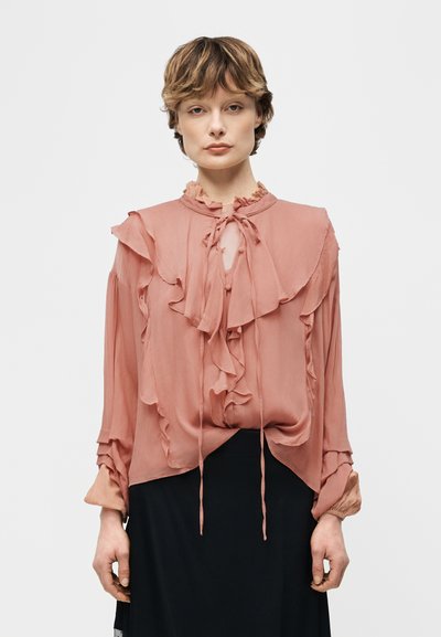 Femme aux cheveux courts portant un chemisier rose poudré à volants avec manches longues et une encolure goutte d'eau, associé à une jupe noire.