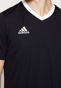 Černé sportovní tričko s výstřihem do V a krátkými rukávy. Má bílé detaily na límci a vyšívané bílé logo Adidas na prsou.