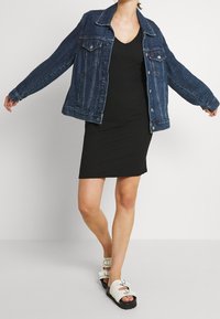 Giubbotto di jeans blu scuro, con colletto classico, chiusura a bottoni e due tasche sul petto, indossato sopra un vestito nero aderente e sandali bianchi.