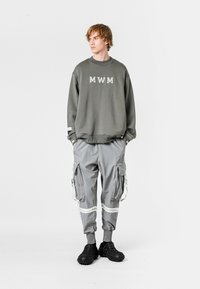 Sudadera gris con la inscripción "MWM", combinada con pantalones cargo gris claro que cuentan con múltiples bolsillos y acentos blancos, llevada con zapatos negros.