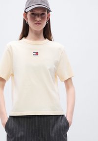 Μπεζ βαμβακερό μπλουζάκι με στρογγυλή λαιμόκοψη και κοντά μανίκια, με ταμπέλα με λογότυπο σε κόκκινο και μπλε. Συνδυασμένο με γκρι παντελόνι με ρίγες.