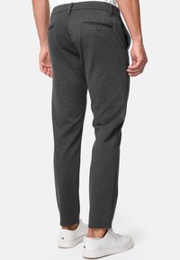 Homme portant un pantalon slim gris foncé et des baskets blanches, vu de dos sur un fond blanc uni.