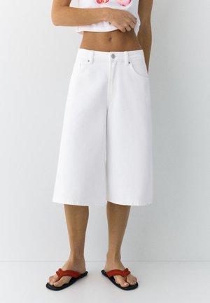 Kvinde iført hvide knælange vide shorts, cropped hvid top og røde klipklappere, står mod en ensfarvet hvid baggrund.