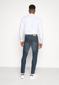 Skinny-fit dark blue jeans with subtle fading, paired with a light blue striped shirt and white sneakers. Features a branded tag on the back.