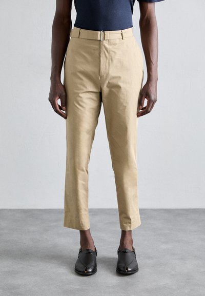 Khaki bawełniane spodnie o prostym kroju, z paskiem z metalową klamrą i skróconą długością, zestawione z czarnymi butami typu slip-on.