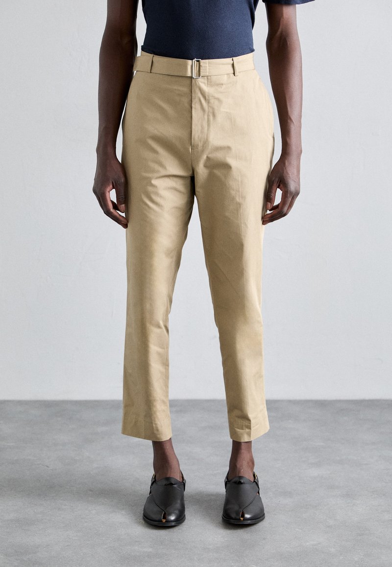 Pantalons en coton kaki à coupe droite, dotés d'une ceinture avec une boucle métallique et d'une longueur coupée, associés à des chaussures noires à enfiler.