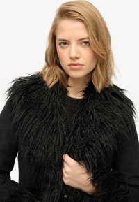 Sort faux fur frakke med en shaggy krave, der har en tekstureret overflade. Materialet ser blødt ud og er parret med en strikket sweater under.