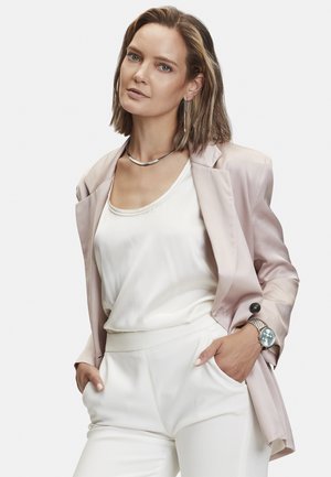 Helle pinke Satinhüfte über einem weißen Seidentop und weißen, maßgeschneiderten Hosen. Mit einer silbernen Halskette und einer silbernen Uhr mit blauem Ziffernblatt.